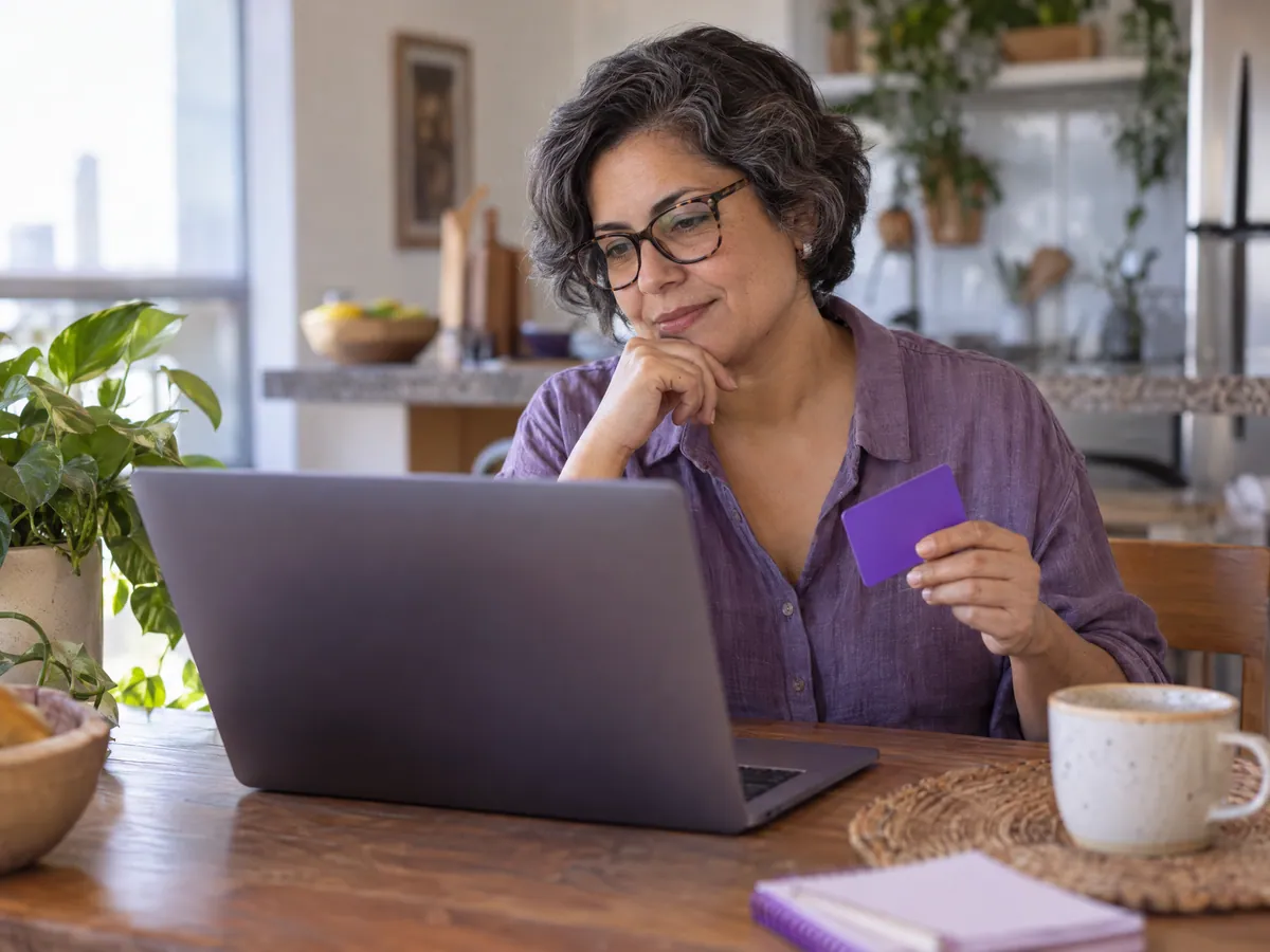 A woman at her kitchen table holds a card next to her laptop